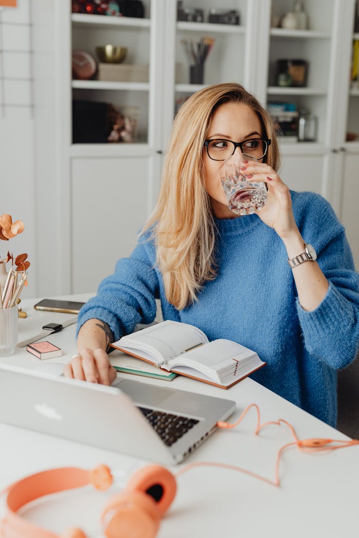 Blonde woman working from home with a laptop, notebook, and drink, embracing remote work lifestyle.